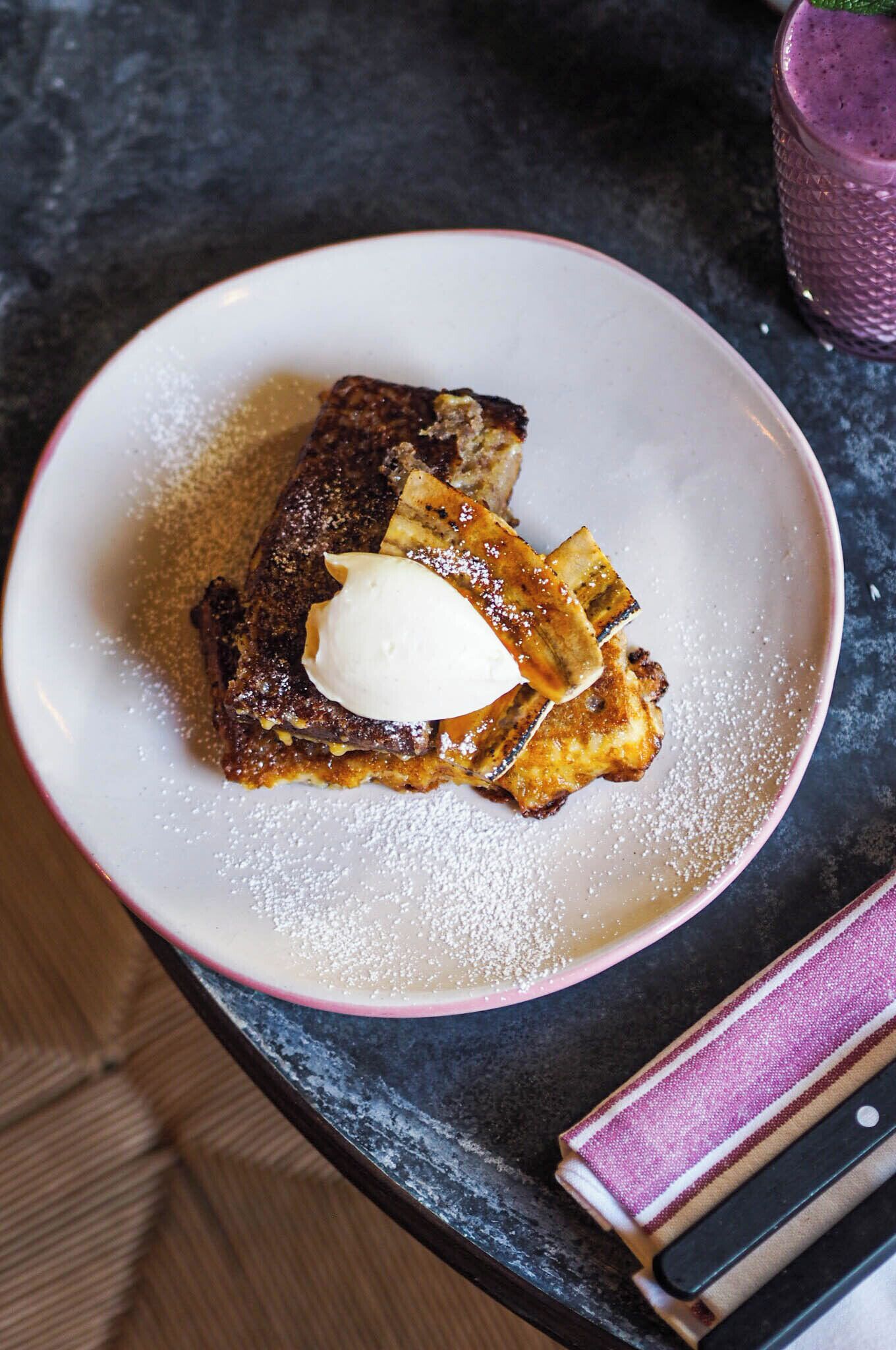 Une assiette de pain perdu, surmontée d’une cuillère de crème fouettée et saupoudrée de sucre glace, posée sur une surface sombre.