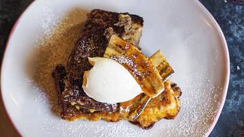 Une assiette de pain perdu, surmontée d’une cuillère de crème fouettée et saupoudrée de sucre glace, posée sur une surface sombre.