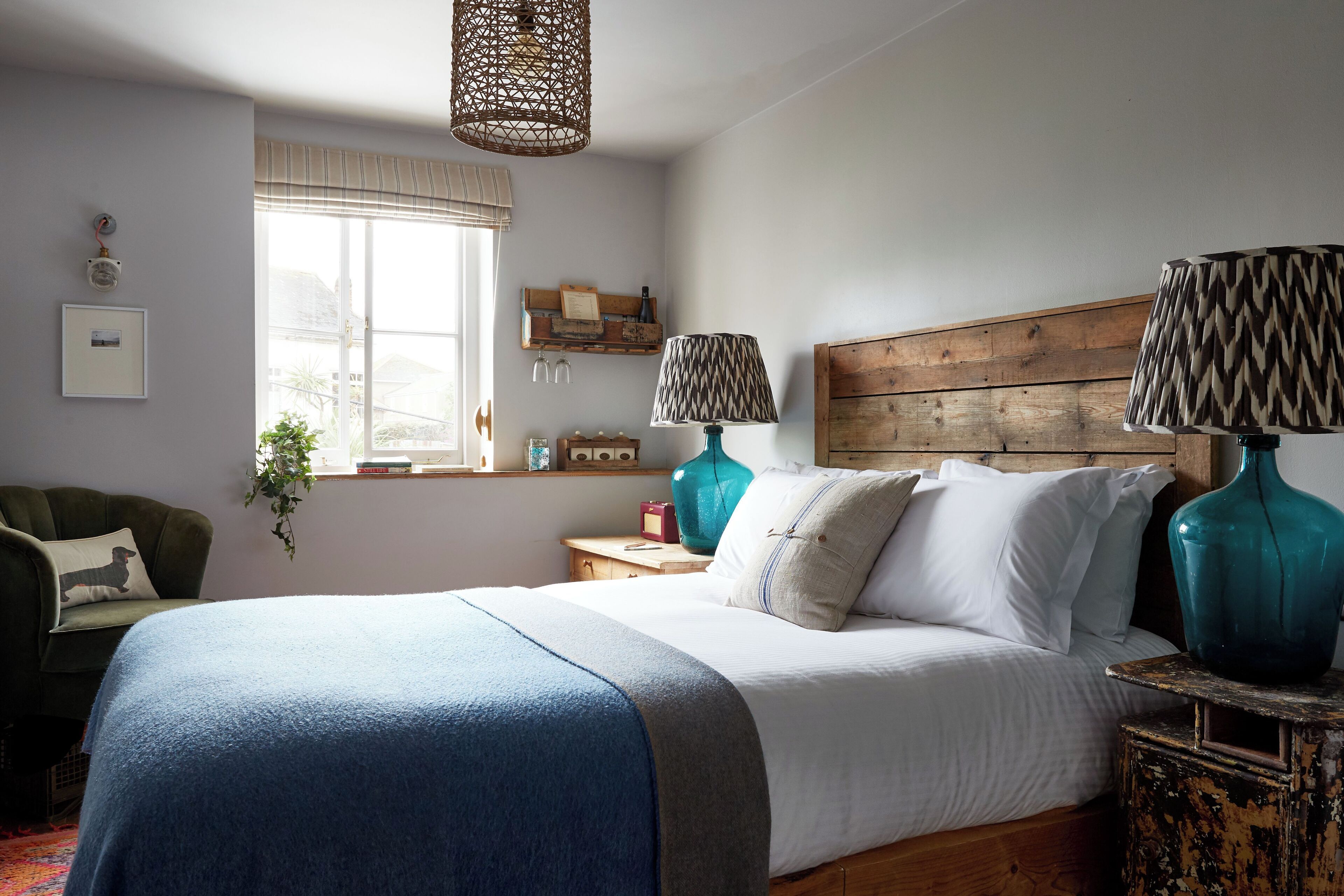 A bedroom with a large bed, two bedside lamps, a wooden headboard, a window with blinds, a green armchair, and a small table with a vase.