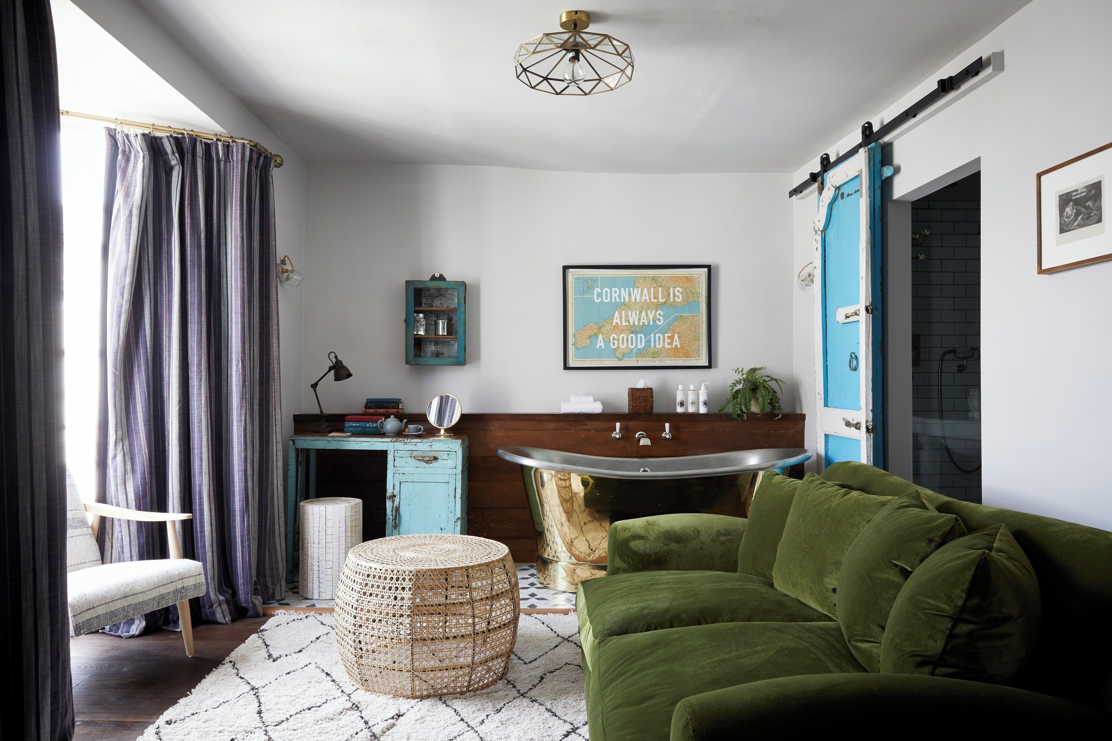 A cozy living room with a green sofa, a woven ottoman, a blue cabinet, and a large window with curtains.