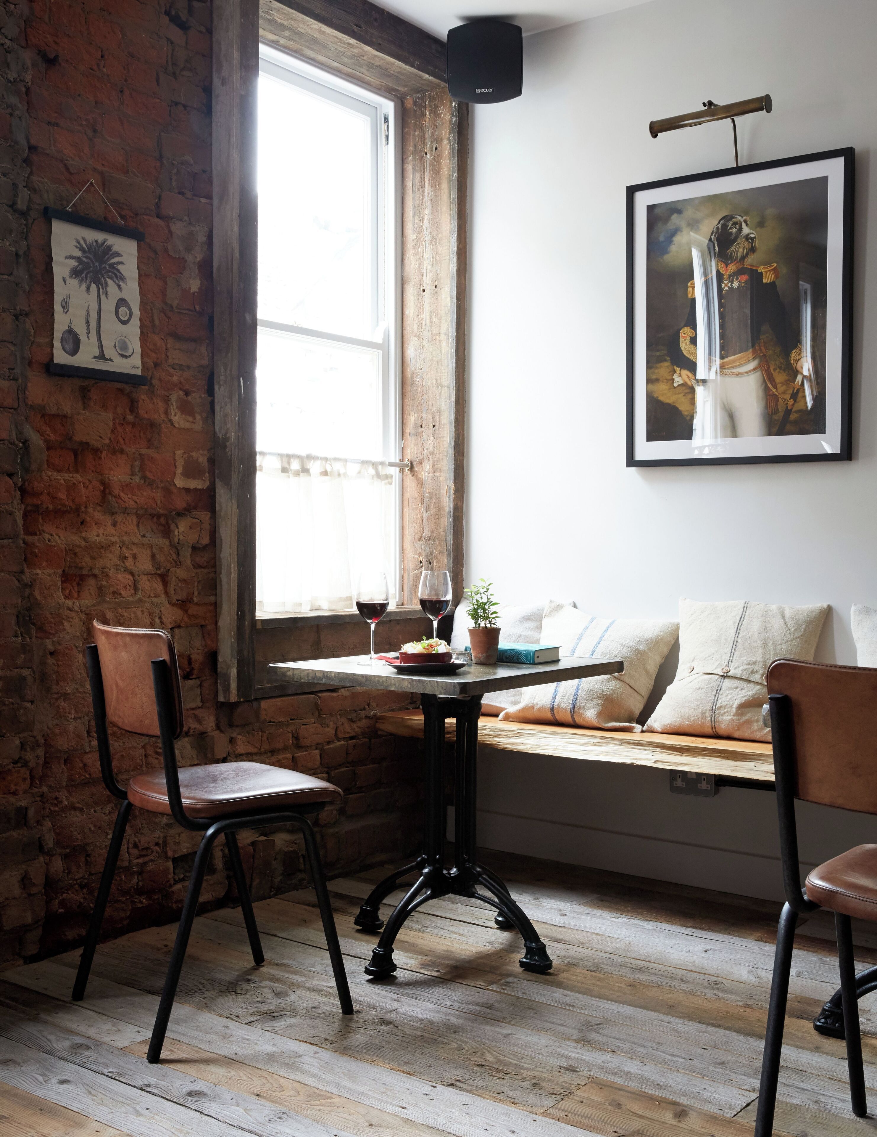Une pièce chaleureuse avec un mur en briques, un plancher en bois, une fenêtre avec des rideaux, un tableau encadré et une table agrémentée d’une plante et de verres à vin.