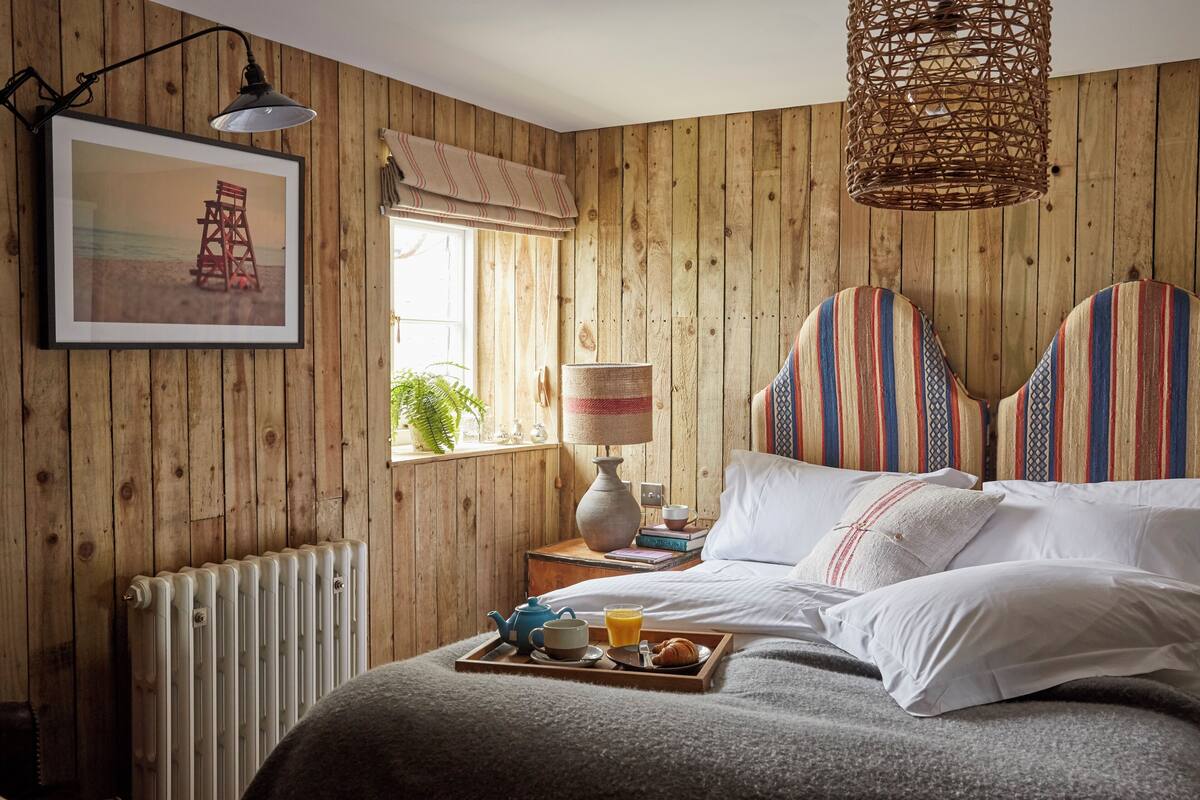A cozy bedroom with wooden walls, a bed with striped pillows, a bedside table with a lamp, and a tray with breakfast items.