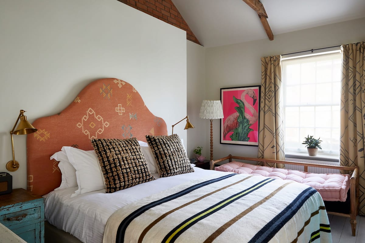 A bedroom with a bed, a nightstand, a lamp, a window with curtains, and a wall with a painting.
