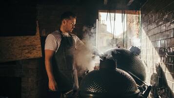 A person standing next to a large smoker with smoke visible, in a room with brick walls and a window.