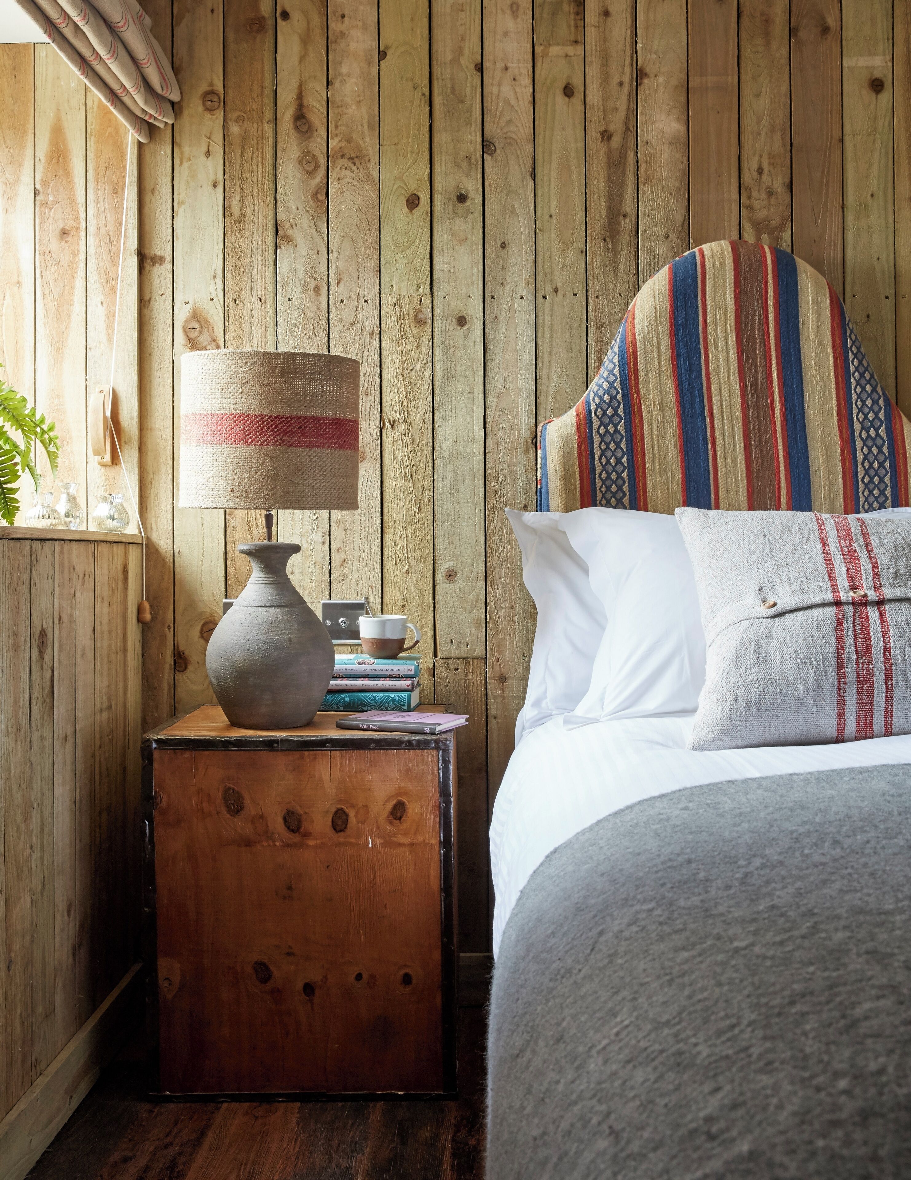 Una stanza in legno con un letto, un comodino con una lampada, libri e una piccola pianta.