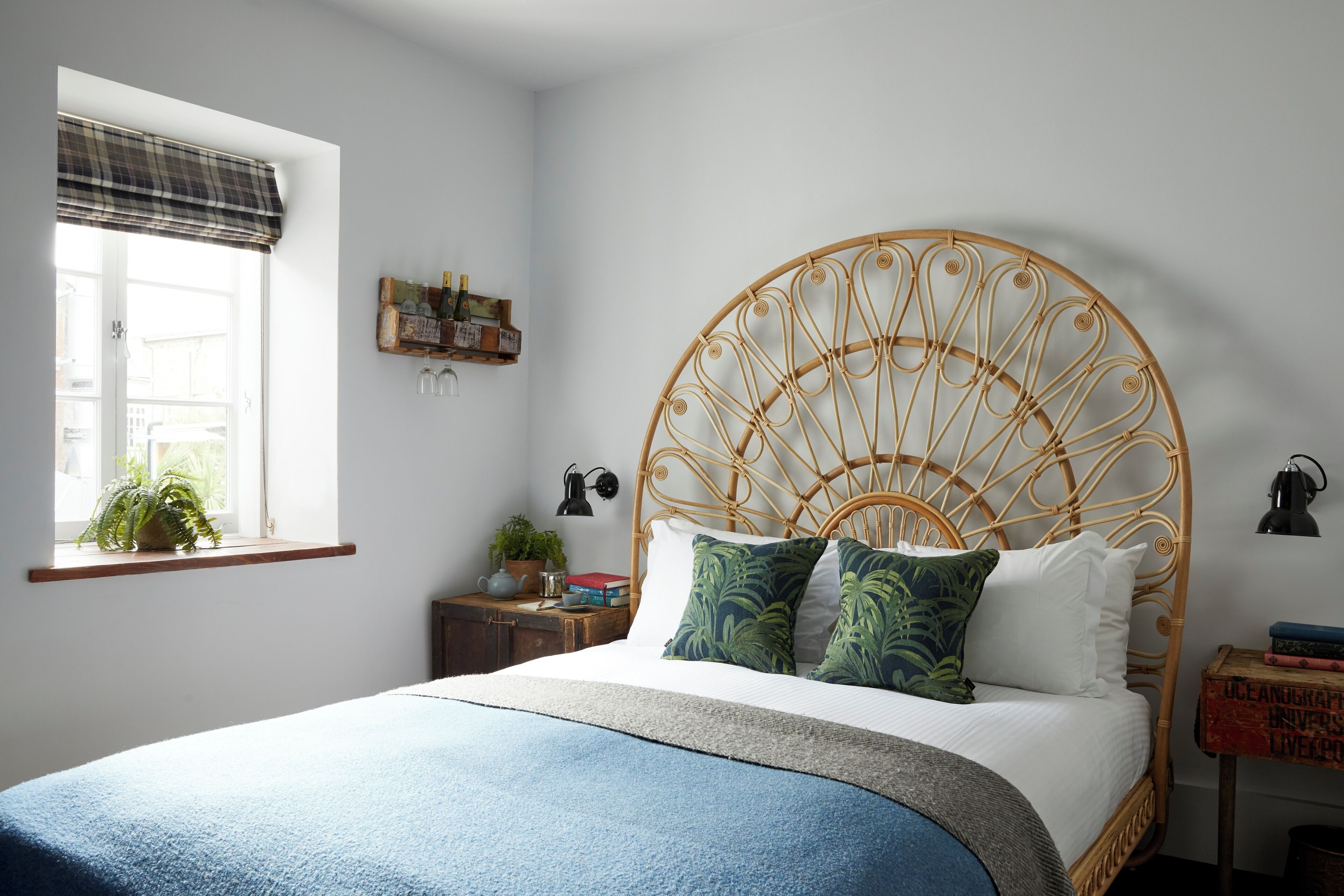 A bedroom with a large bed, a window with a view, a bedside table with a lamp, and a small shelf with books and a plant.