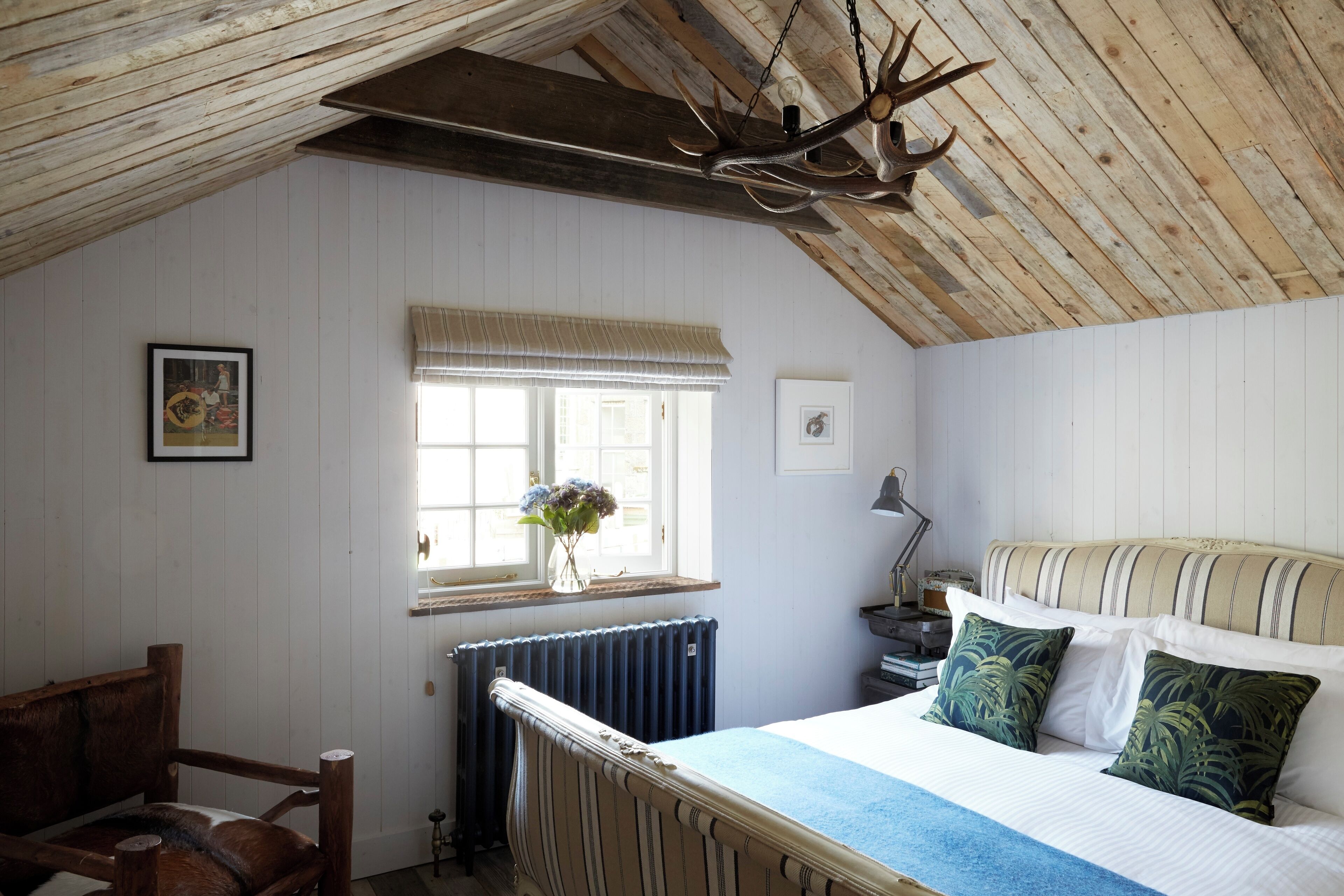 Un dormitorio con techo de madera, una cama con ropa de cama a rayas, un radiador, una ventana con vistas y una silla de madera.