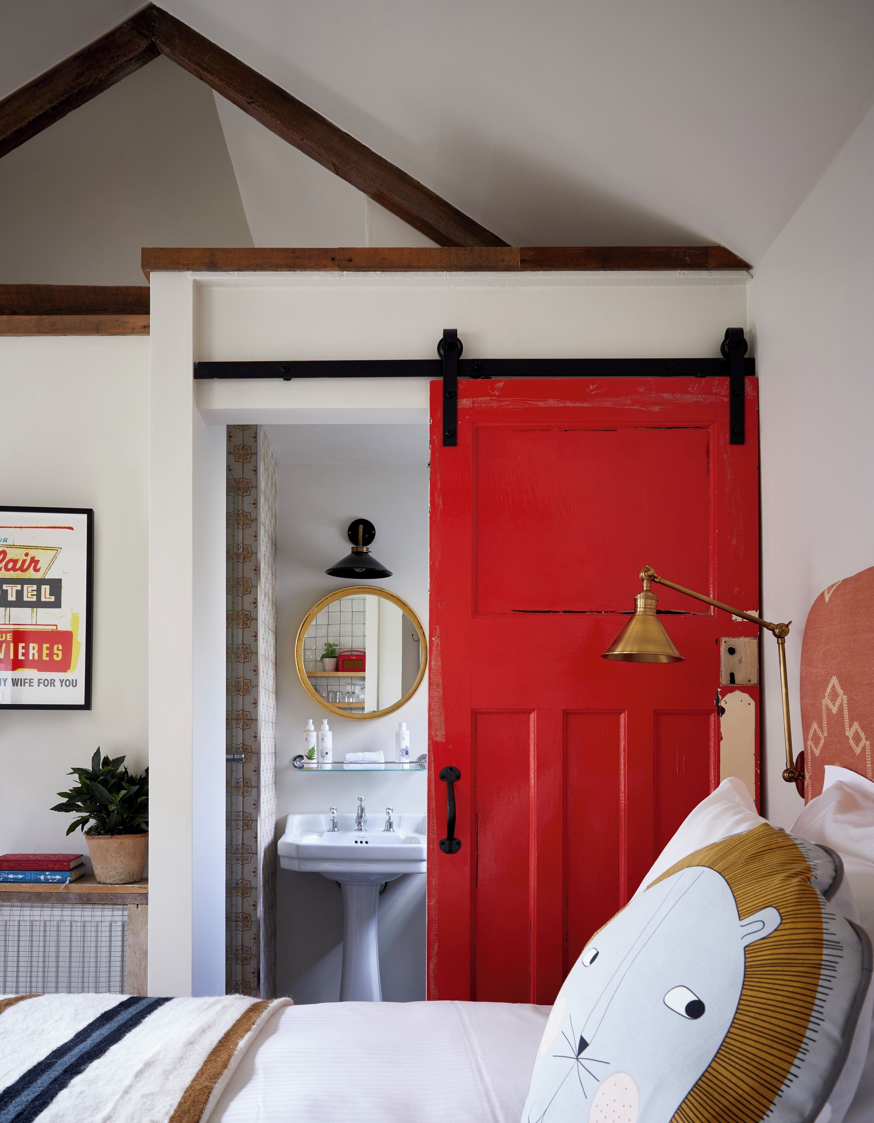 Um quarto com porta vermelha, cama com manta listrada em branco e marrom, luminária de cabeceira, espelho fixado na parede e um pequeno vaso de planta.