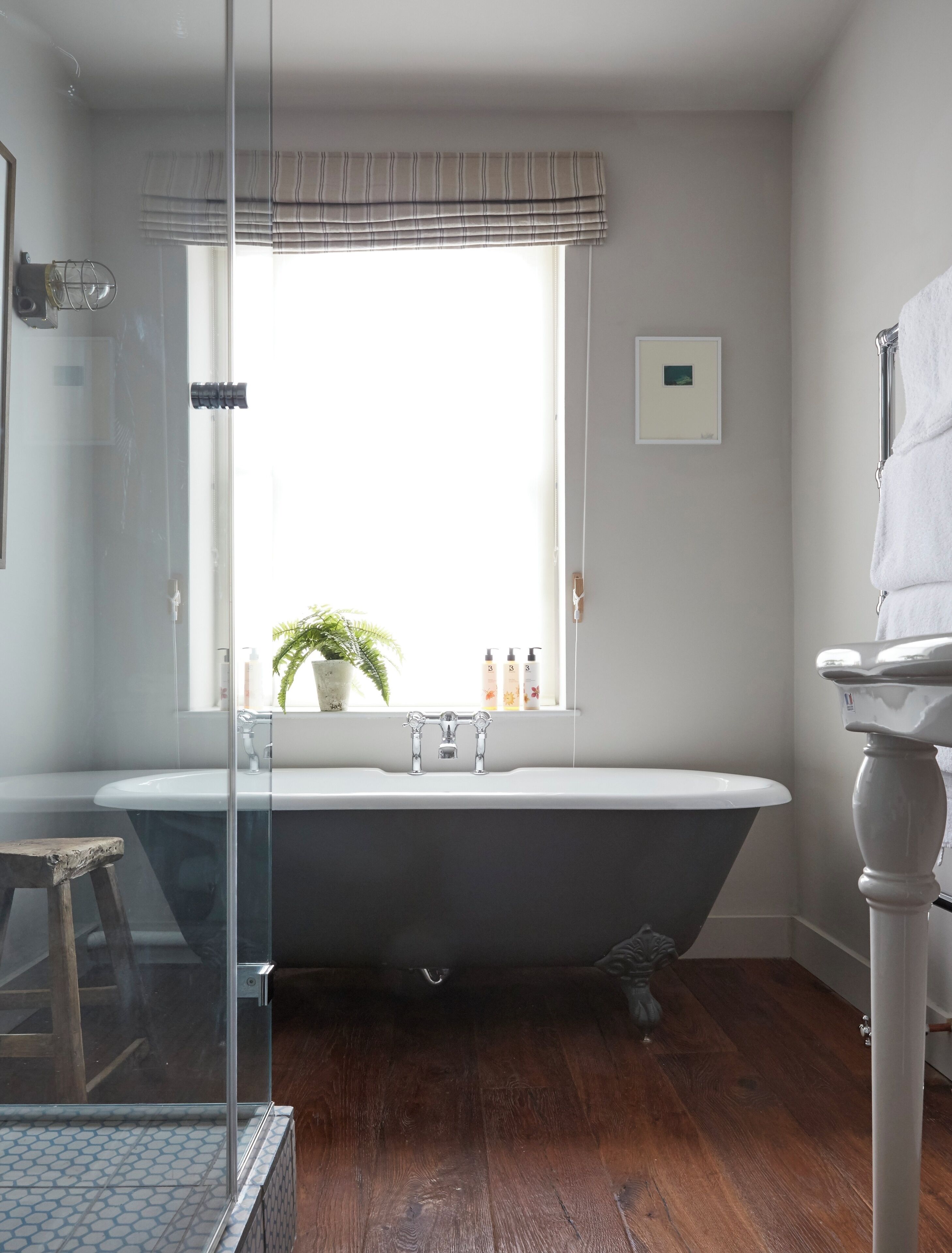 A bathroom with a freestanding bathtub, a glass shower enclosure, a wooden stool, and a window with blinds.