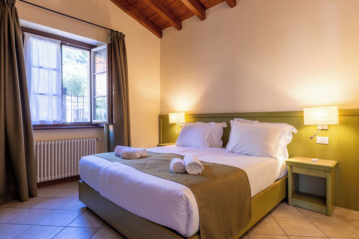 A neatly made bed with white linens and a brown throw, two bedside tables with lamps, a window with curtains, and a radiator.