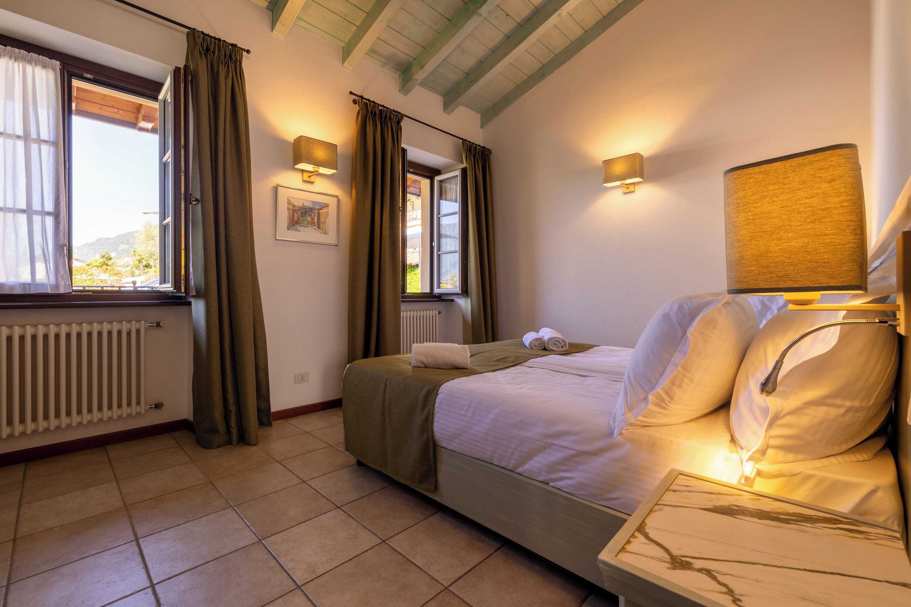 Habitación de hotel con cama, dos ventanas con cortinas, un radiador y lámparas fijadas a la pared.