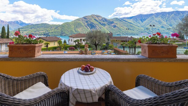 A terrace with wicker chairs and a table, overlooking a lake and mountains. - Hotel Villa Edy (Tremezzina)