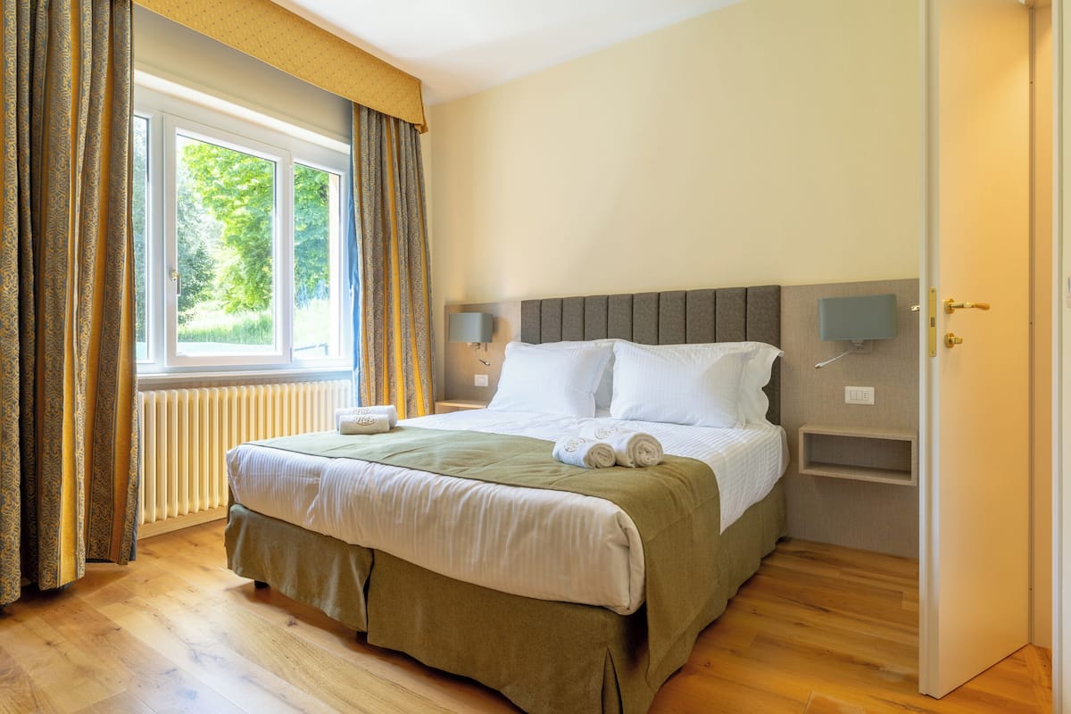 A neatly made bed with a green bed skirt, a bedside table with a lamp, and a window with curtains.