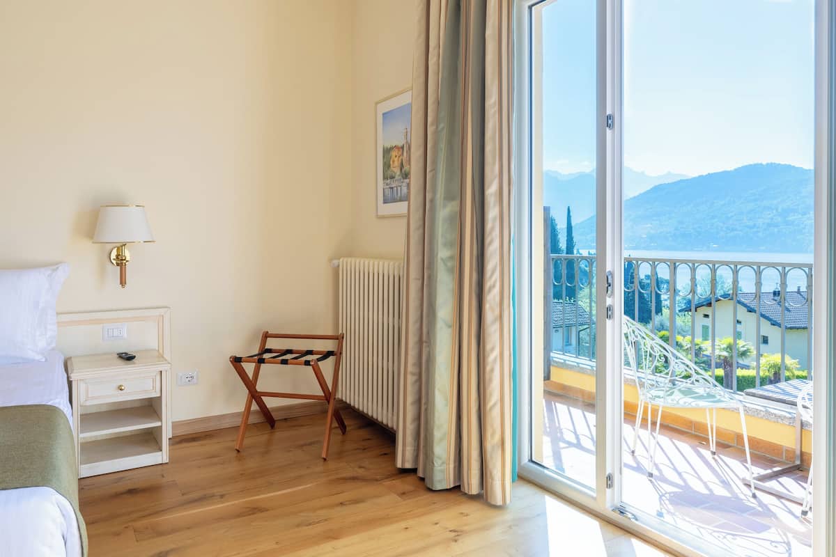 A hotel room with a wooden floor, a bed, a nightstand with a lamp, a chair, a radiator, and a large sliding glass door leading to a balcony with a view of a mountain and buildings.