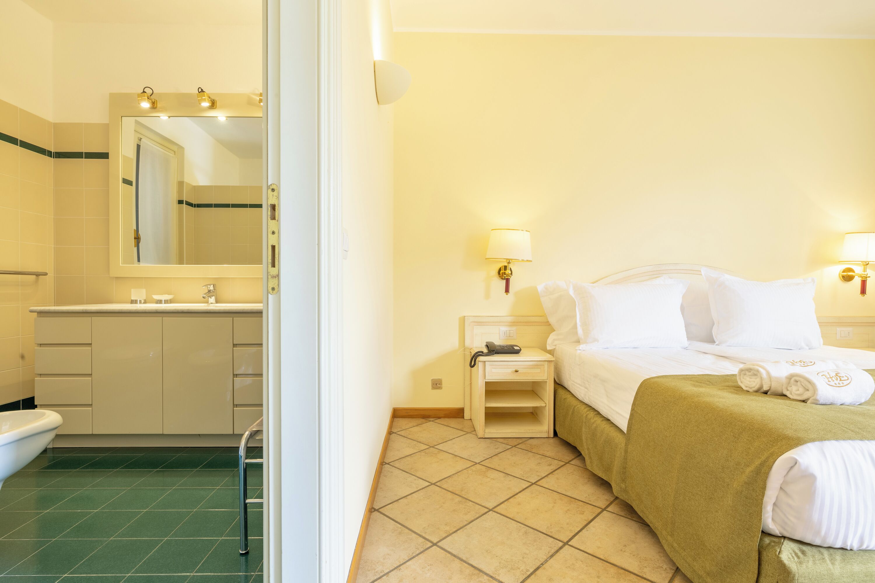 Ein Hotelzimmer mit Bad, einem Bett mit weißen Laken, einer grünen Tagesdecke und einem Nachttisch mit Lampe.