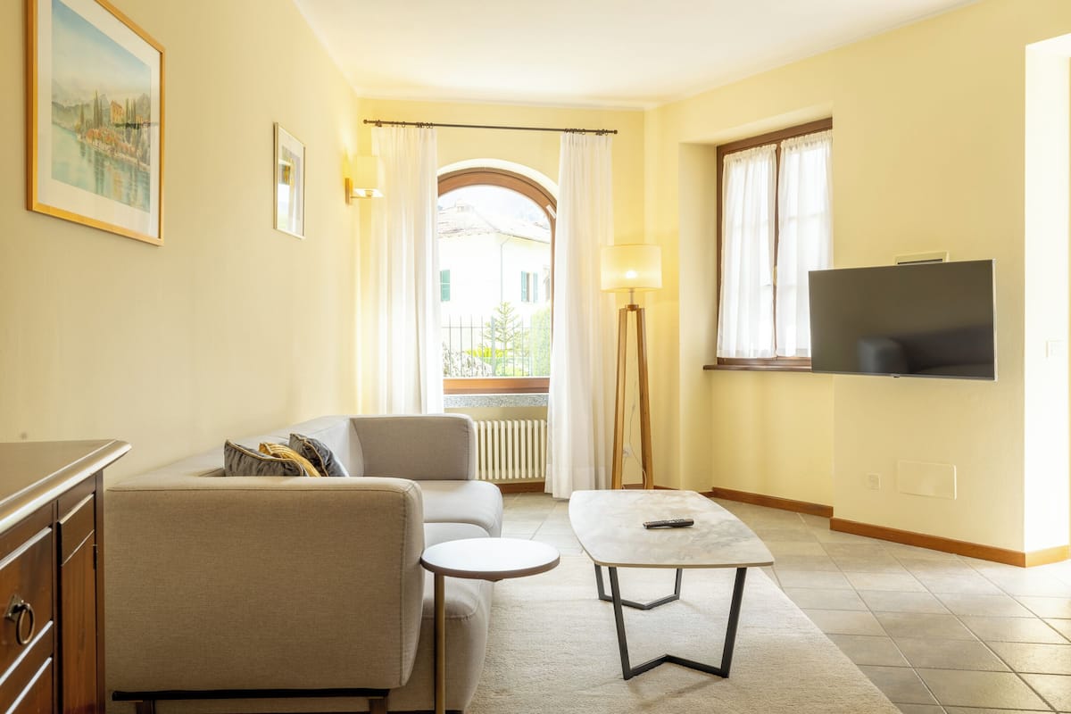 A living room with a sofa, a small round table, a TV mounted on the wall, and a window with curtains.