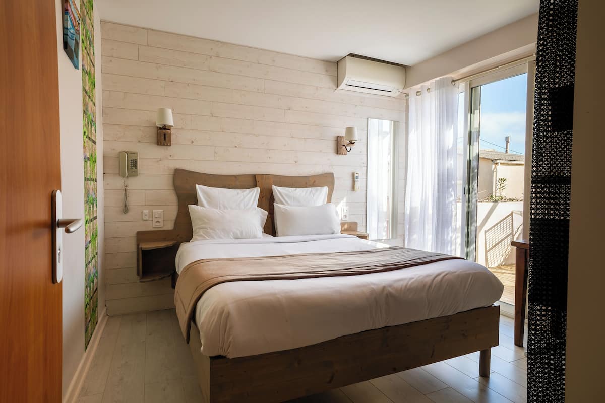 Un dormitorio con cama de madera, ropa de cama blanca, una mesita de noche, teléfono en la pared y una ventana con cortinas.