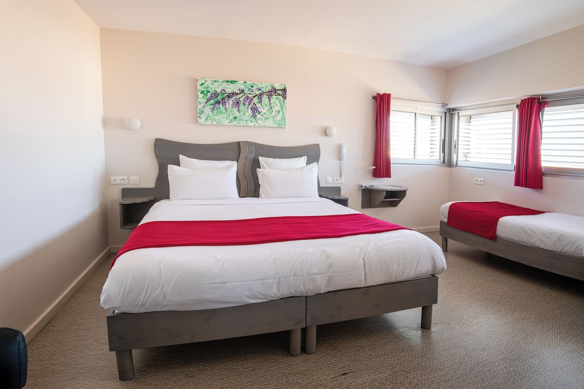 Una habitación de hotel con una cama grande, dos mesitas de noche, una lámpara de noche, una ventana con cortinas rojas y una obra de arte colgada en la pared.