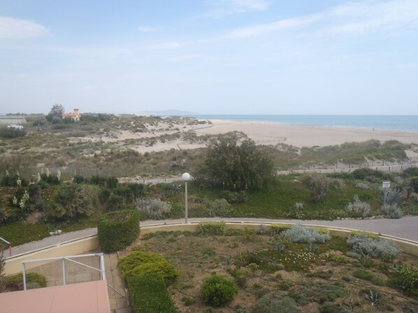 A coastal landscape with a sandy beach, a fenced area with vegetation, and a building with a red roof in the distance. - Hôtel Les Dunes (Marseillan)