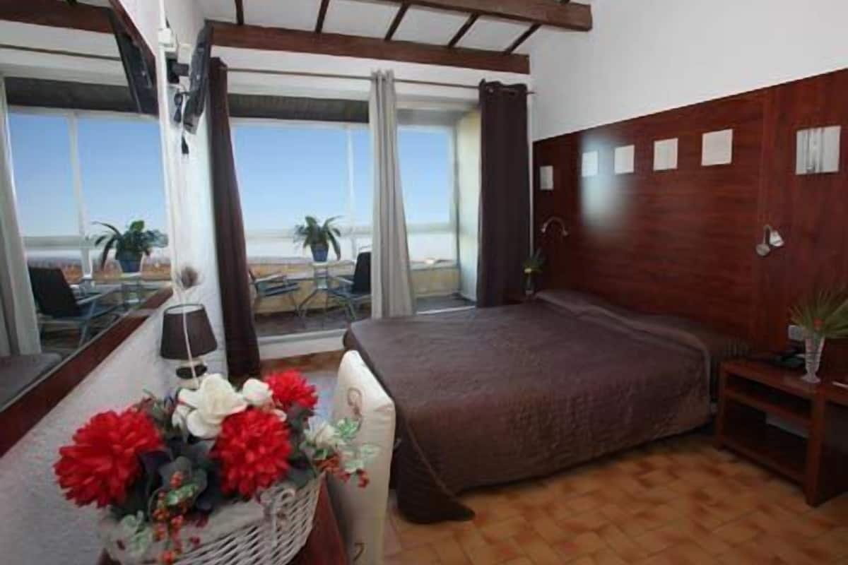 A bedroom with a large bed, a window with a view of the beach, a desk with a lamp, and a basket of flowers.