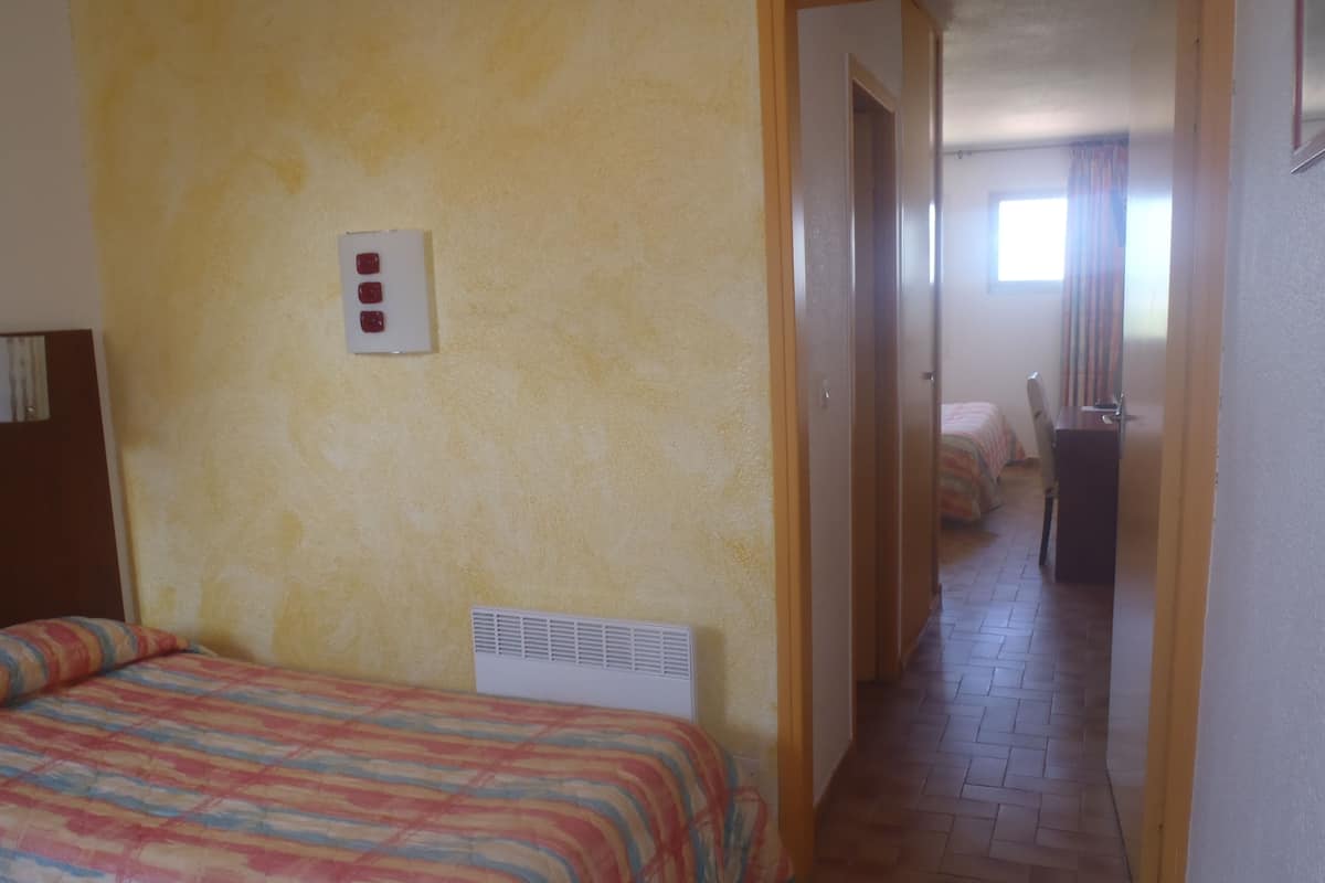 A hotel room with a bed, a heater, a wall-mounted light switch, and a view into the adjacent room.