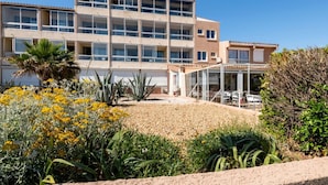 Ein mehrstöckiges Hotel mit Balkonen, einer überdachten Außenbereich-Sitzgelegenheit und einem angelegten Garten mit verschiedenen Pflanzen und Blumen.
