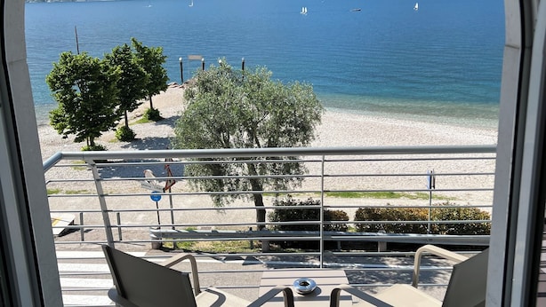A balcony with two chairs and a table, overlooking a beach and a lake with mountains in the background. - San Remo (Malcesine)