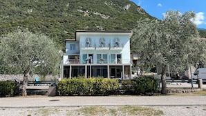 A two-story building with balconies, a gravel driveway, and a mountain backdrop. - San Remo (Malcesine)