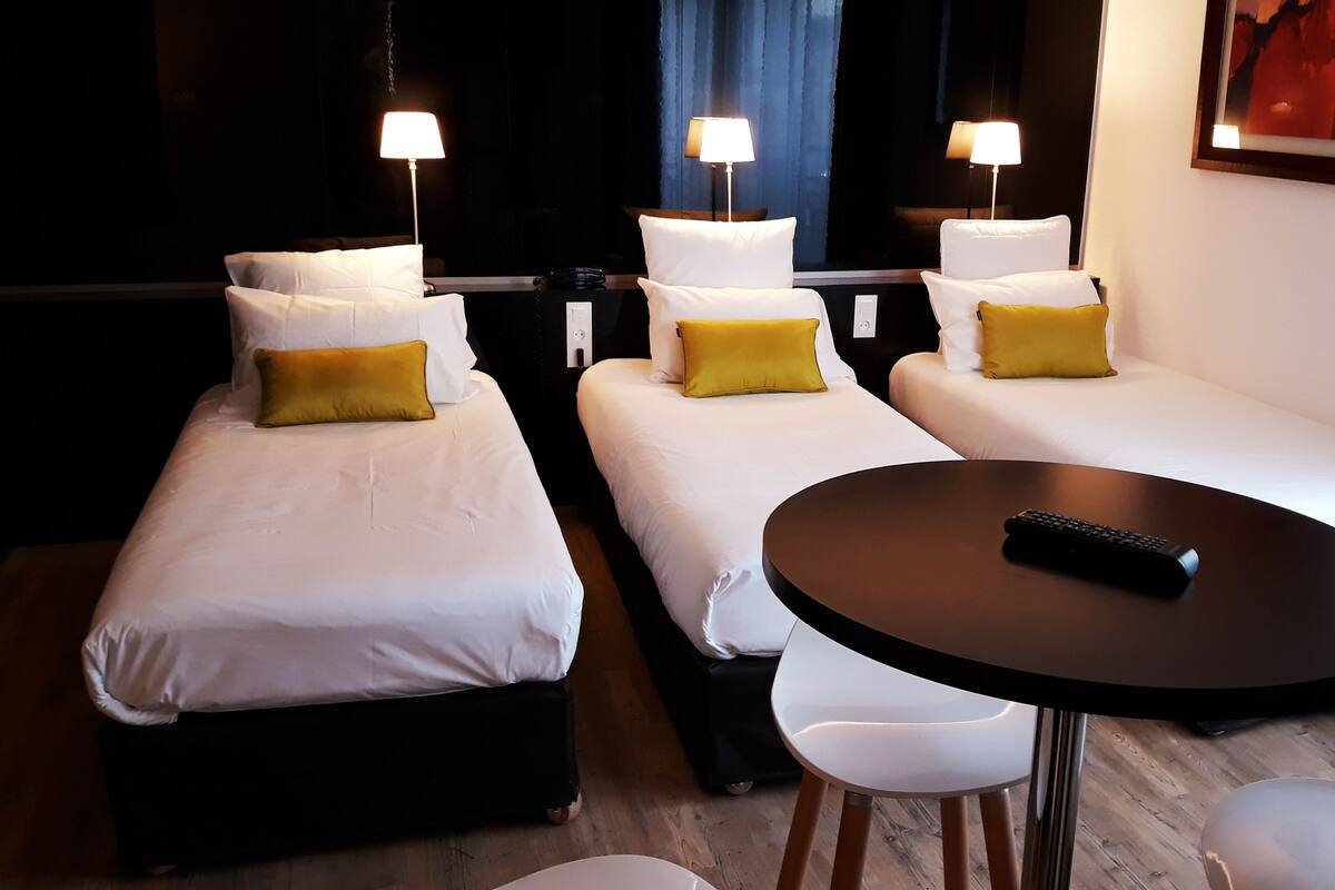 Een hotelkamer met drie eenpersoonsbedden, elk met witte lakens en een geel kussen. Er staat een rond tafel met een afstandsbediening erop.