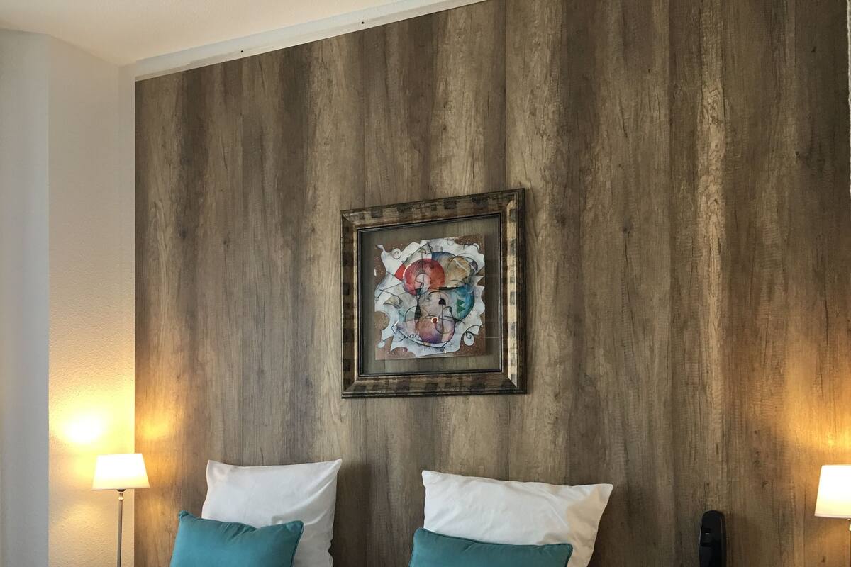 Hotelkamer met een groot bed, twee blauwgroene kussens, een houten wand, een lijst met kunstwerk en een telefoon op het nachtkastje.