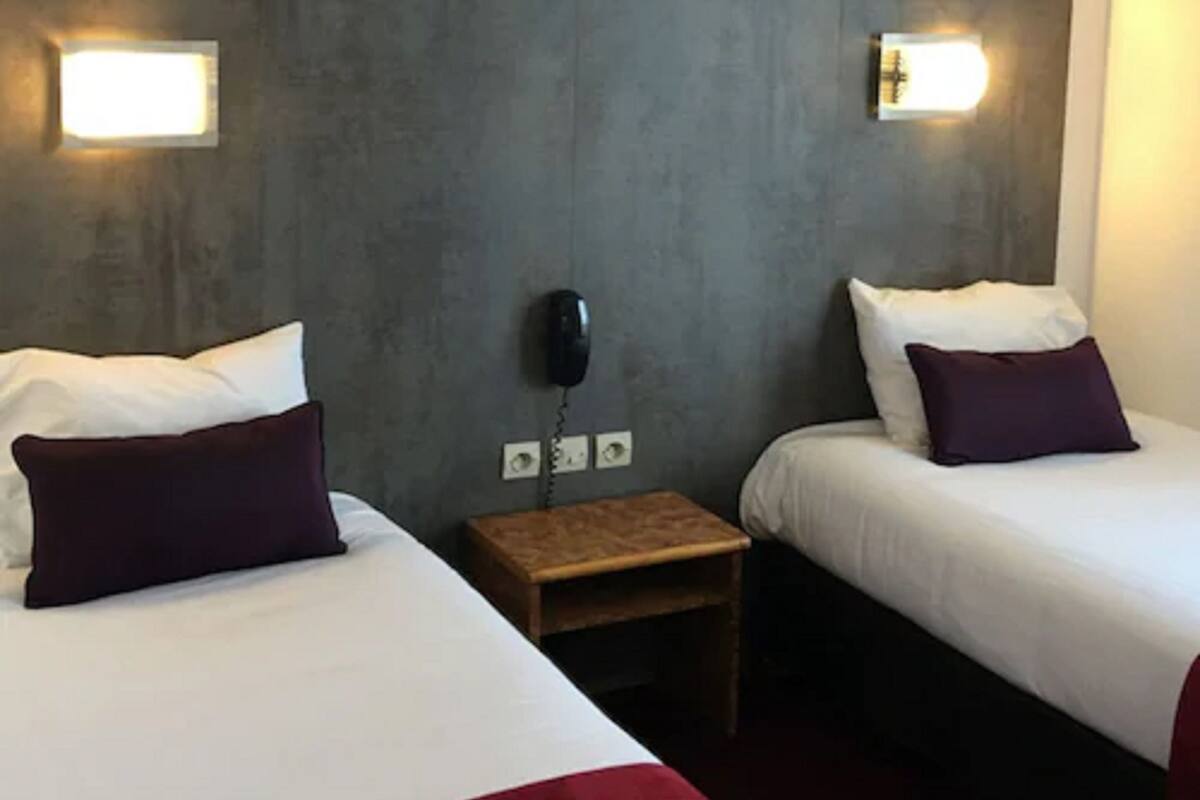 Een hotelkamer met twee bedden, elk voorzien van witte lakens en een donker kussen. Aan weerszijden van de kamer zijn wandlampen bevestigd.