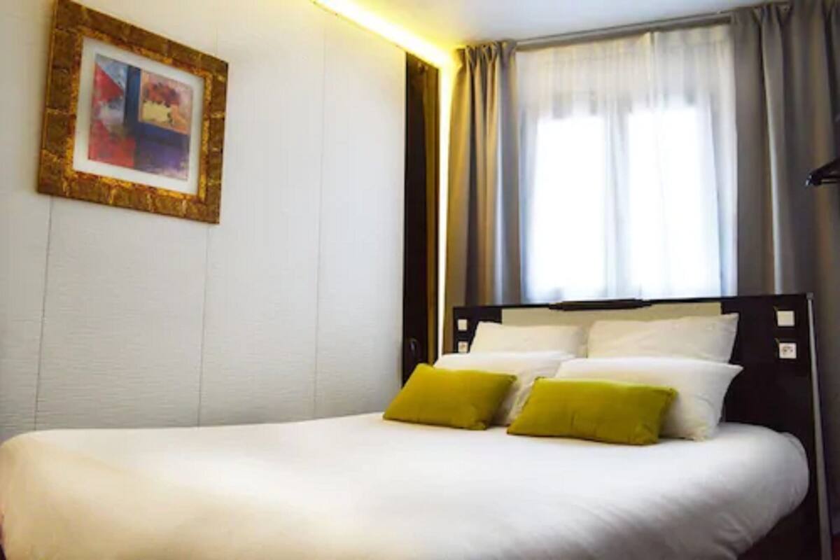 Een netjes opgemaakt bed met witte en gele kussens, een ingelijst beeld aan de muur en een raam met gordijnen.