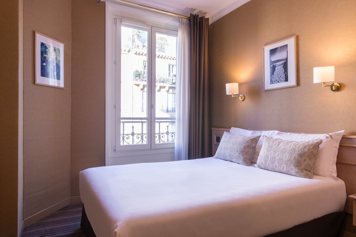 Une chambre d’hôtel avec un grand lit, deux lampes fixées au mur et une fenêtre donnant sur des immeubles.