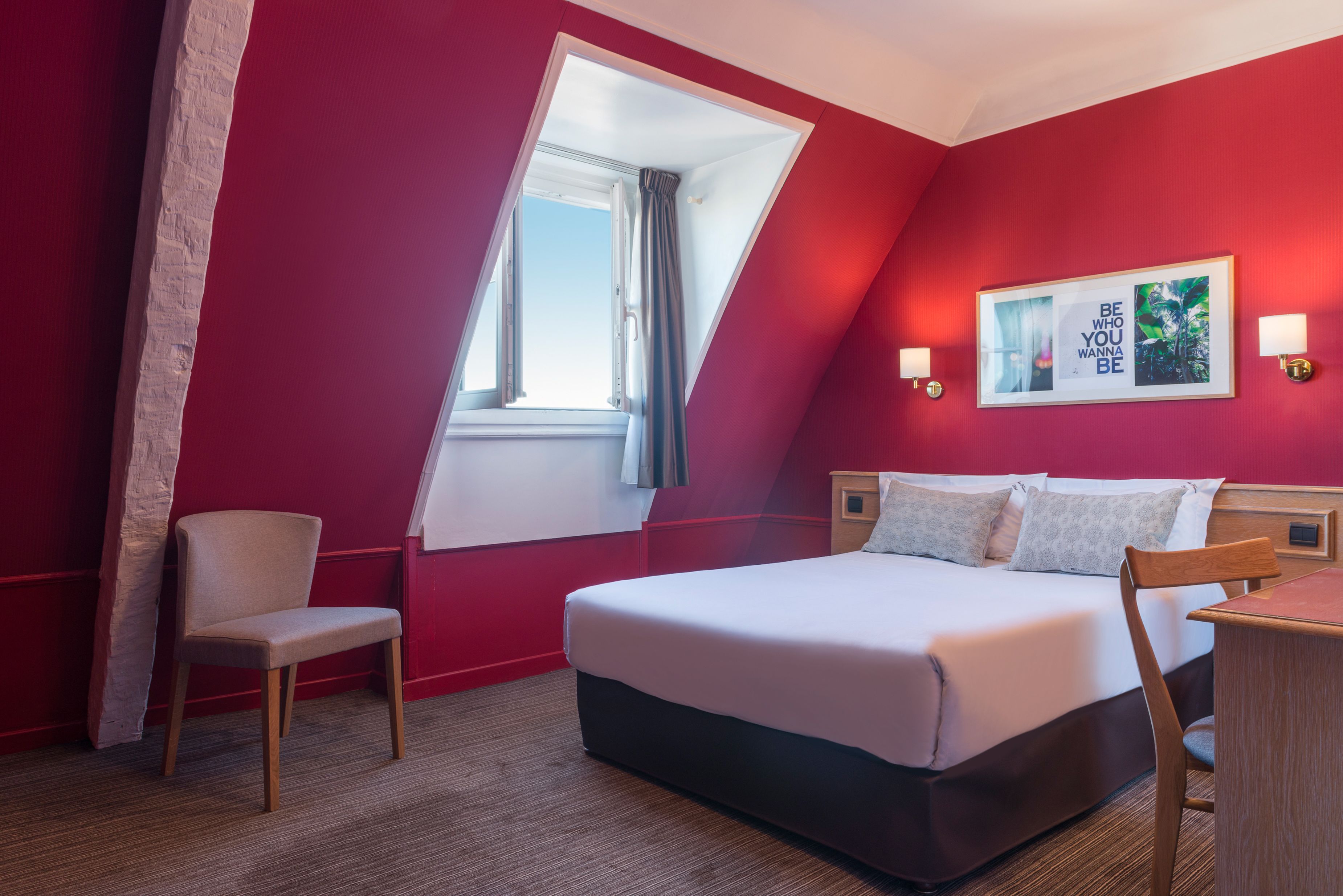 A hotel room with a bed, a chair, a desk, a window, and a wall-mounted picture.