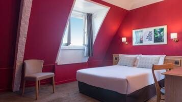 A hotel room with a bed, a chair, a desk, a window, and a wall-mounted picture.
