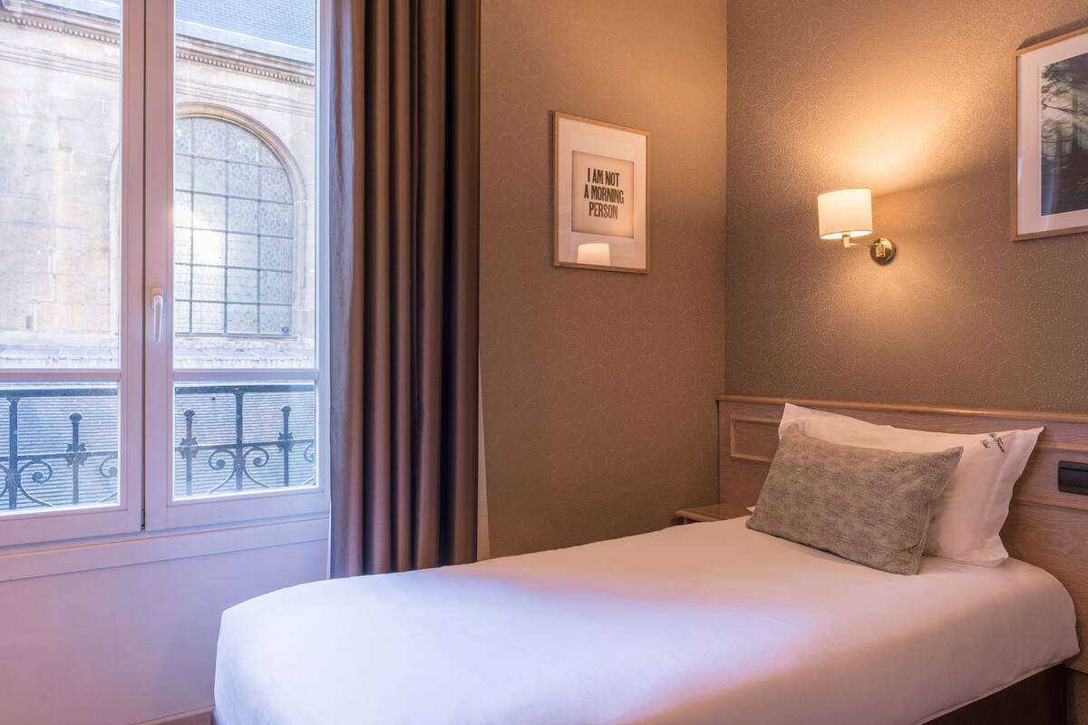 Une chambre d’hôtel avec une grande fenêtre, une citation encadrée au mur, une lampe de chevet et un lit parfaitement fait.