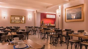 Un comedor con varias mesas y sillas, una cocina al fondo, y cuadros en las paredes.