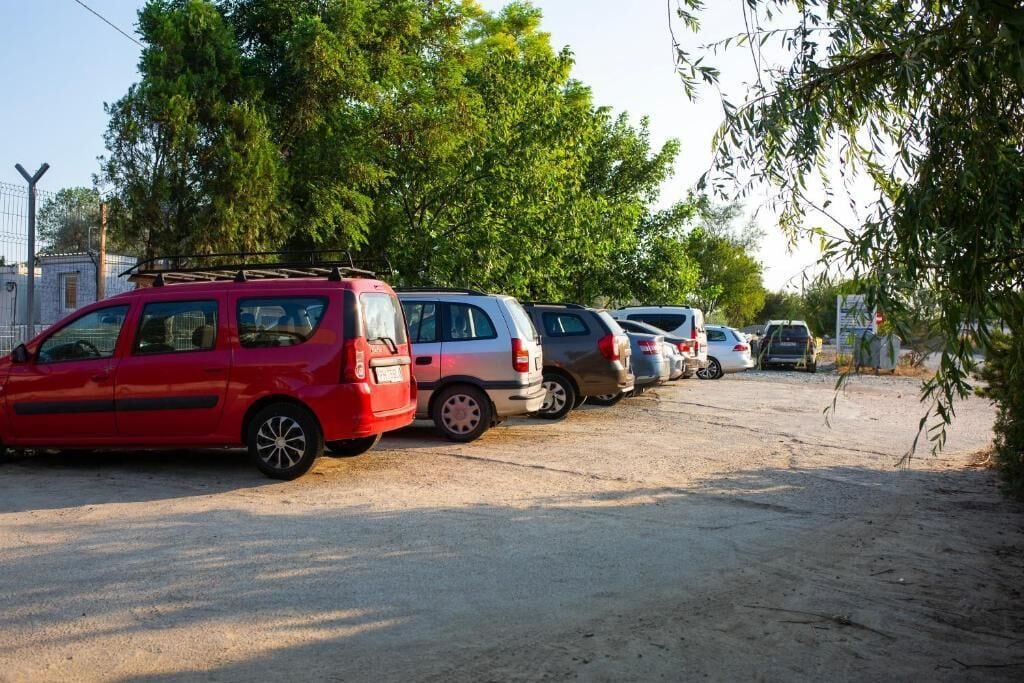 Fila di auto parcheggiate lungo una strada sterrata, con alberi sullo sfondo.
