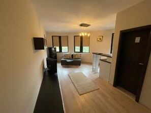 Un pasillo que conduce a una sala de estar con un sofá, una mesa de centro y una alfombra. Hay ventanas con persianas, una puerta y un televisor montado en la pared.