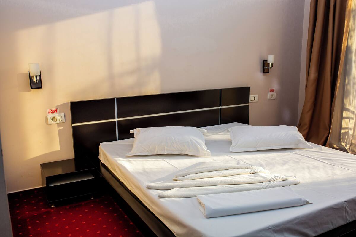 A hotel room with a neatly made bed, a bedside table, a wall-mounted light, and a power outlet.
