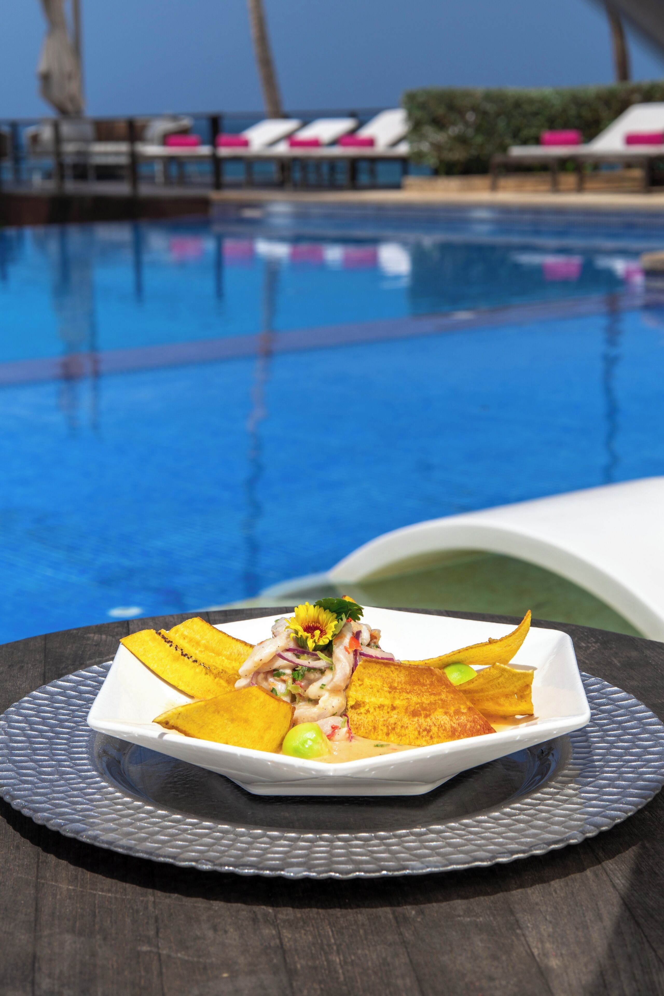 A dining table with a plate of food, a pool in the background, and lounge chairs.