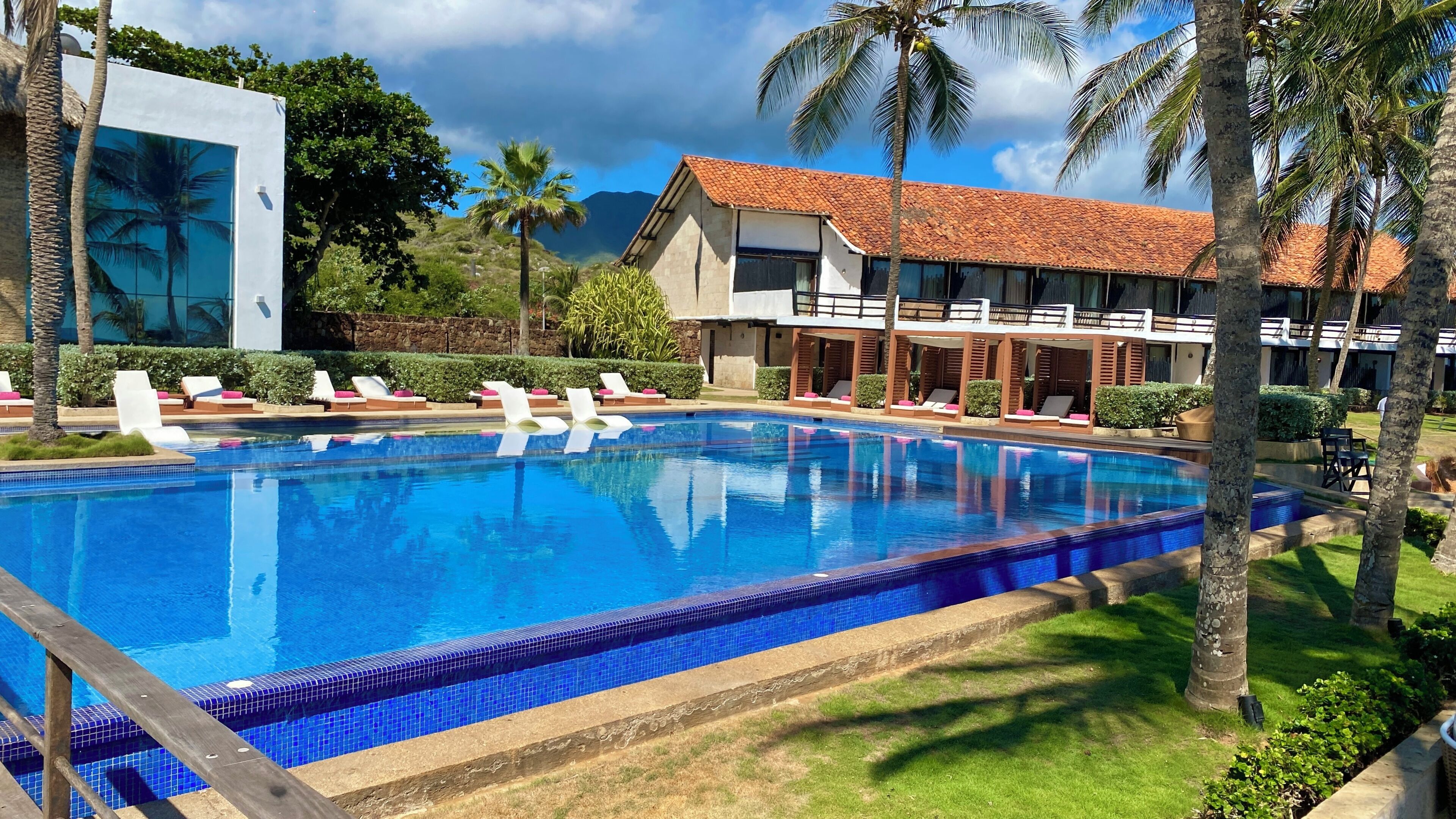 A swimming pool with lounge chairs, a building with a red-tiled roof, and palm trees.