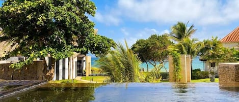 A clear pool with a view of the sea, surrounded by palm trees and a stone wall.
