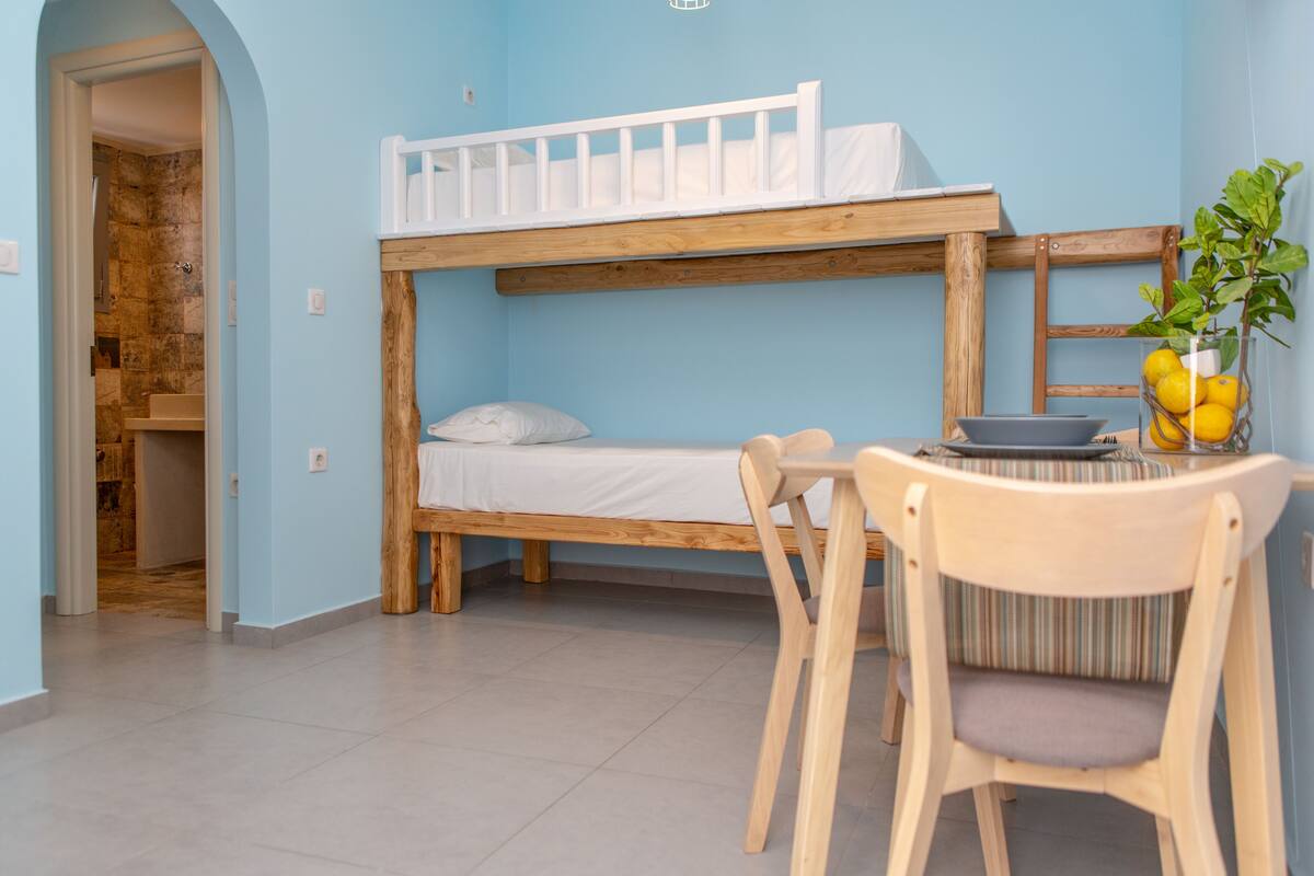 A bunk bed with a wooden frame and white bedding, a small table with a bowl and lemons, and a chair with a striped cushion.