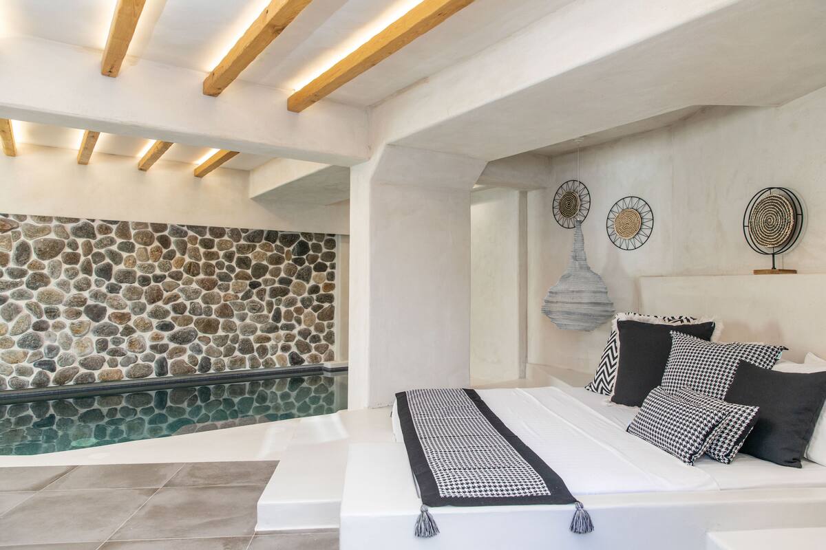 A modern bedroom with a stone wall, a bed with patterned bedding, and a ceiling with exposed beams.