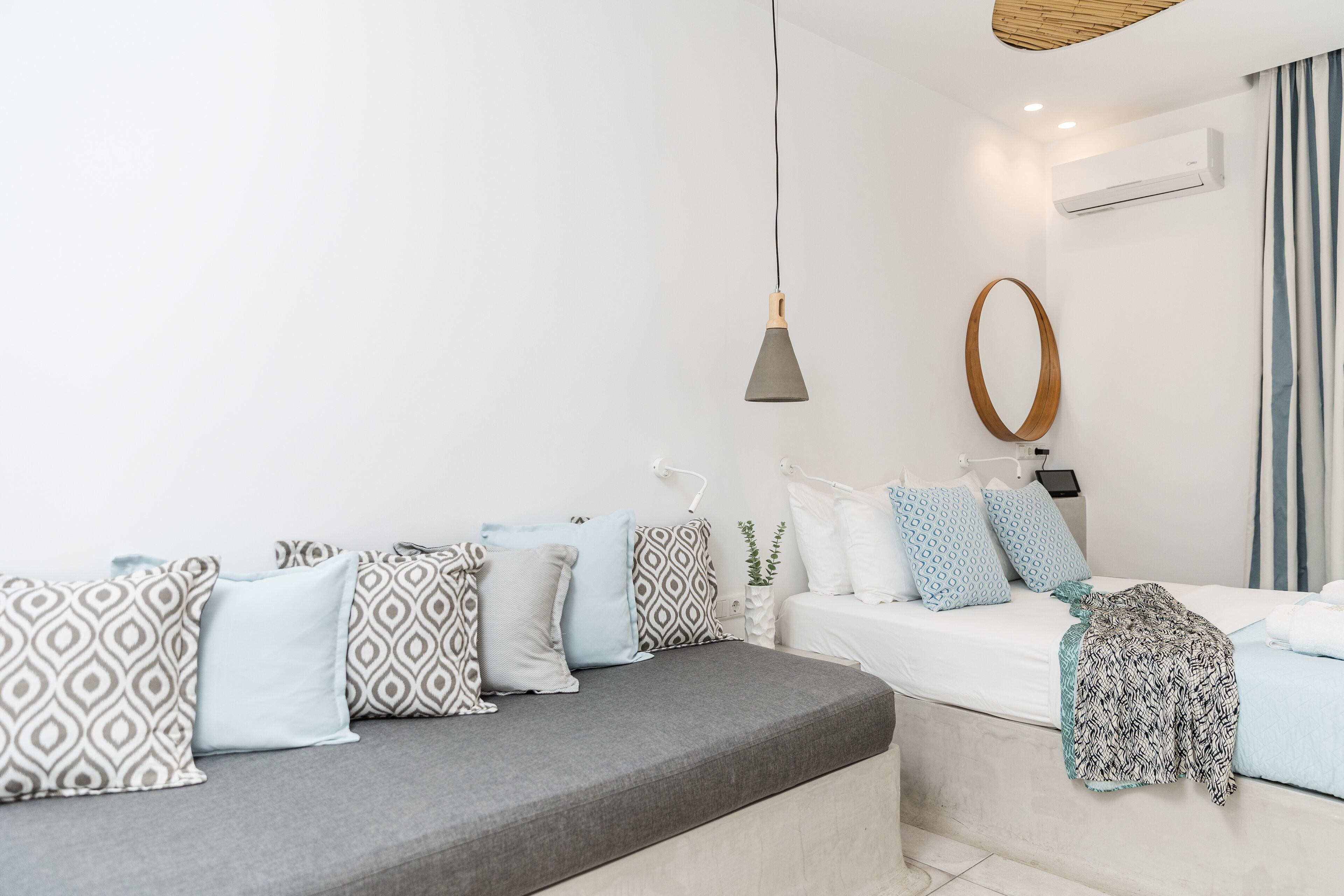 Een moderne hotelkamer met een grijze bank, kussens met een patroon, een spiegel met houten lijst en een hanglamp.