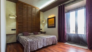 A bedroom with a bed, wooden slat headboard, a wall-mounted light, a window with curtains, and a wooden floor. - Hotel Villa de Setenil (Setenil de las Bodegas)