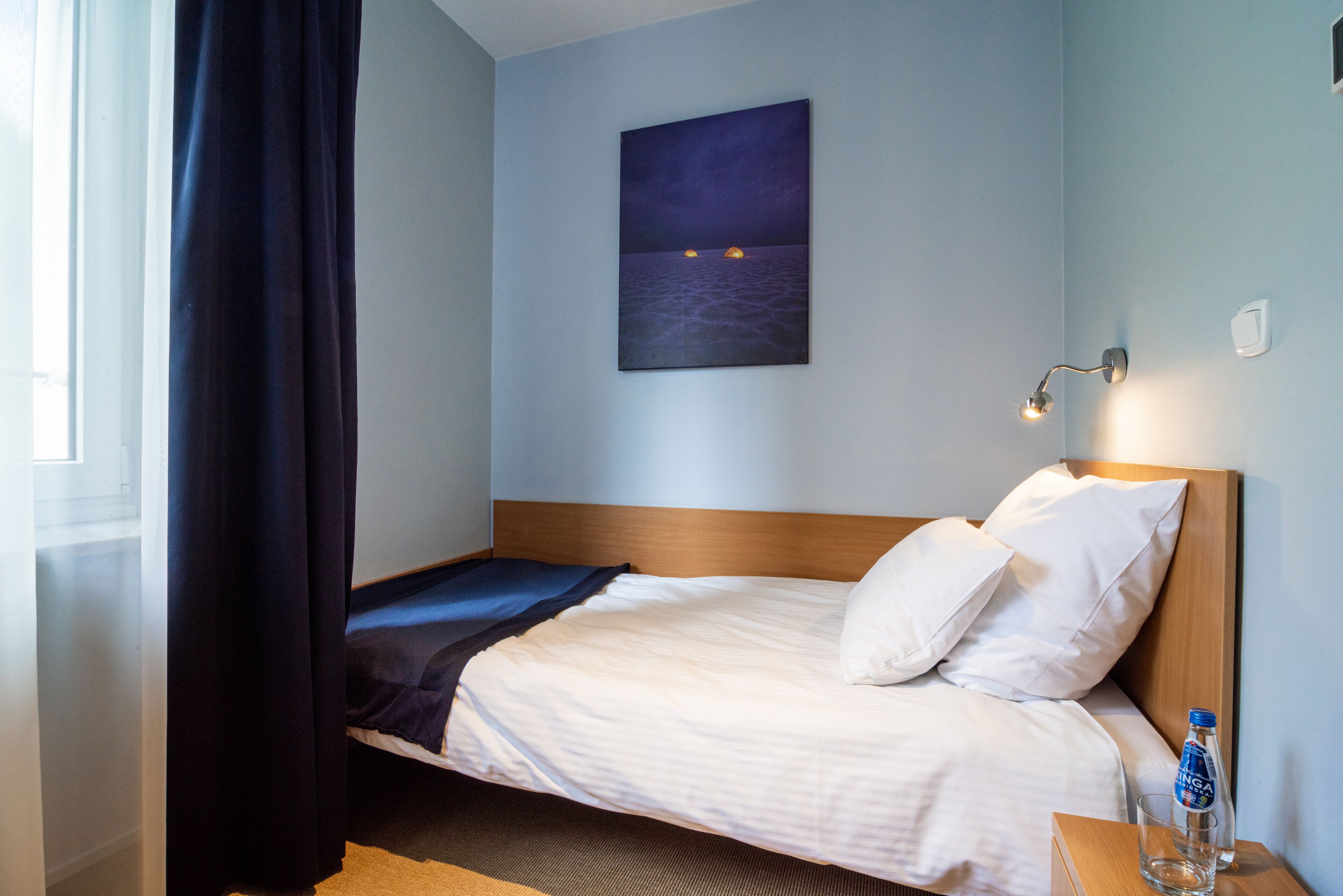A hotel room with a bed, a nightlight, a painting of a night scene, and a bottle of water on the nightstand.