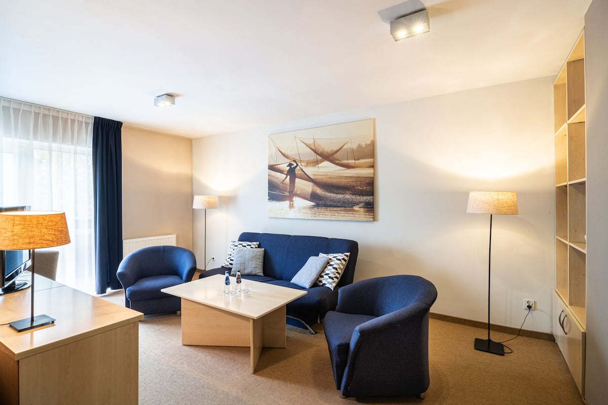 A modern hotel room with a sofa set, a coffee table, a desk with a lamp, and a painting on the wall.