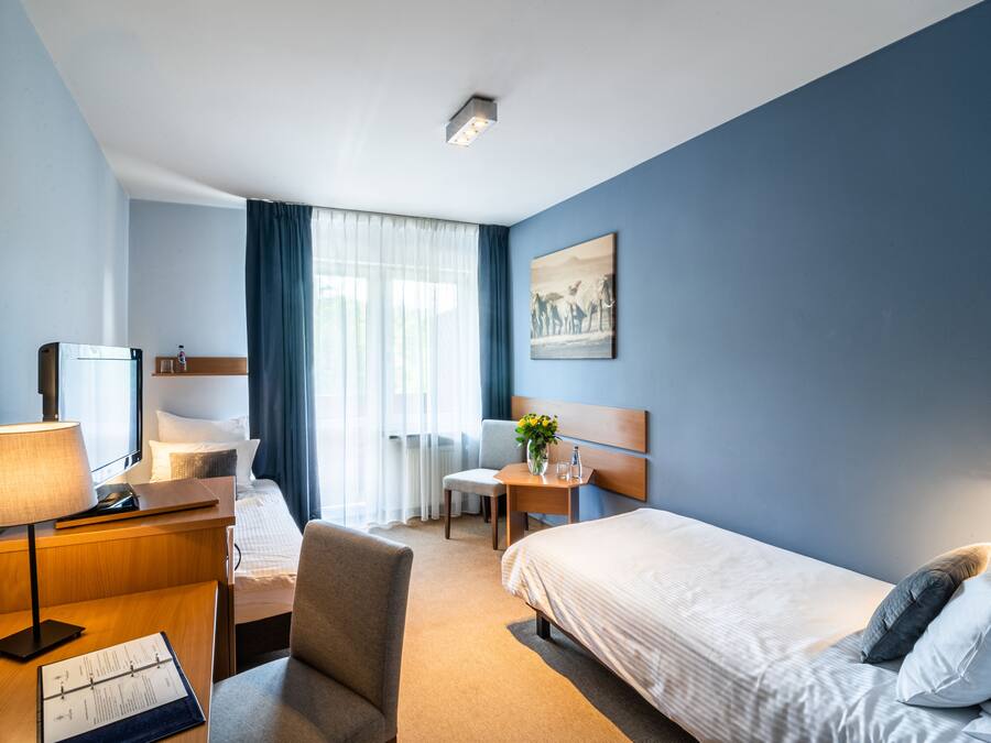A hotel room with a bed, a desk with a lamp, a chair, a small table with a vase of flowers, and a painting on the wall.